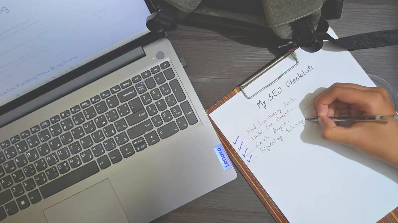 Handwritten SEO checklist on a clipboard next to a laptop, showing steps like finding keywords and requesting indexing.